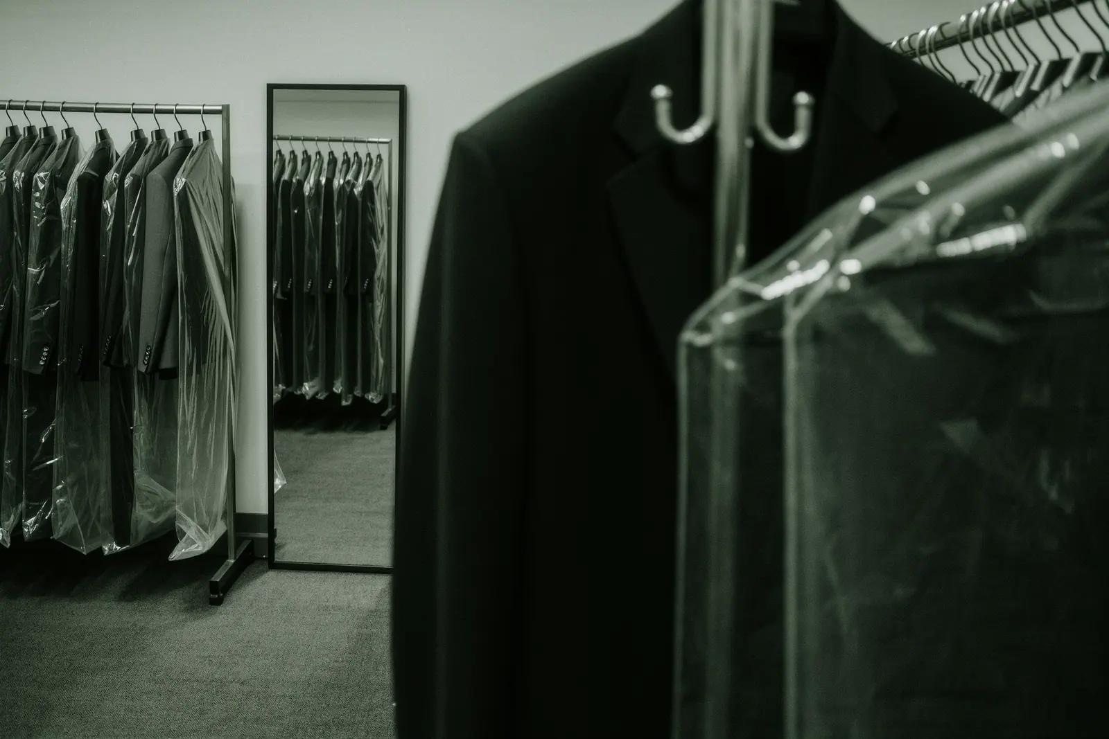 A rail of hire suits and dinner jackets in translucent garment bags beside a plain full-length fitting mirror, the nearest garment bag and hanger hook in the foreground.