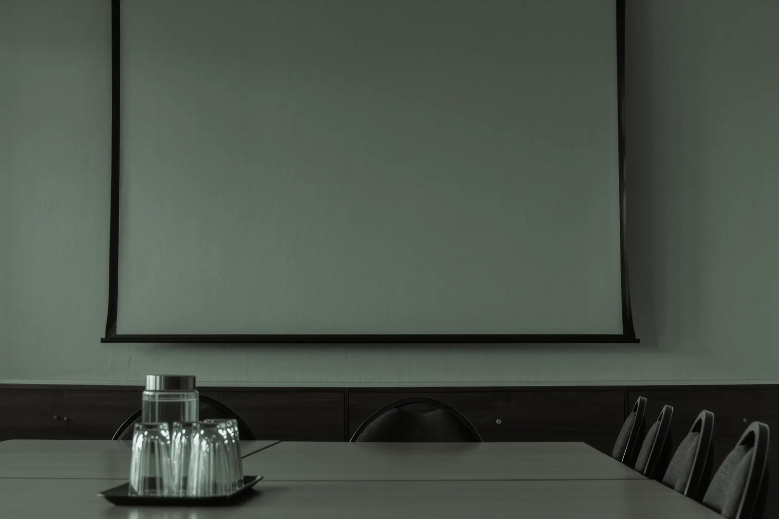A plain hired meeting room with a long table, a water jug and upturned glasses on a tray in the foreground, stacked spare chairs and a blank wall screen behind.
