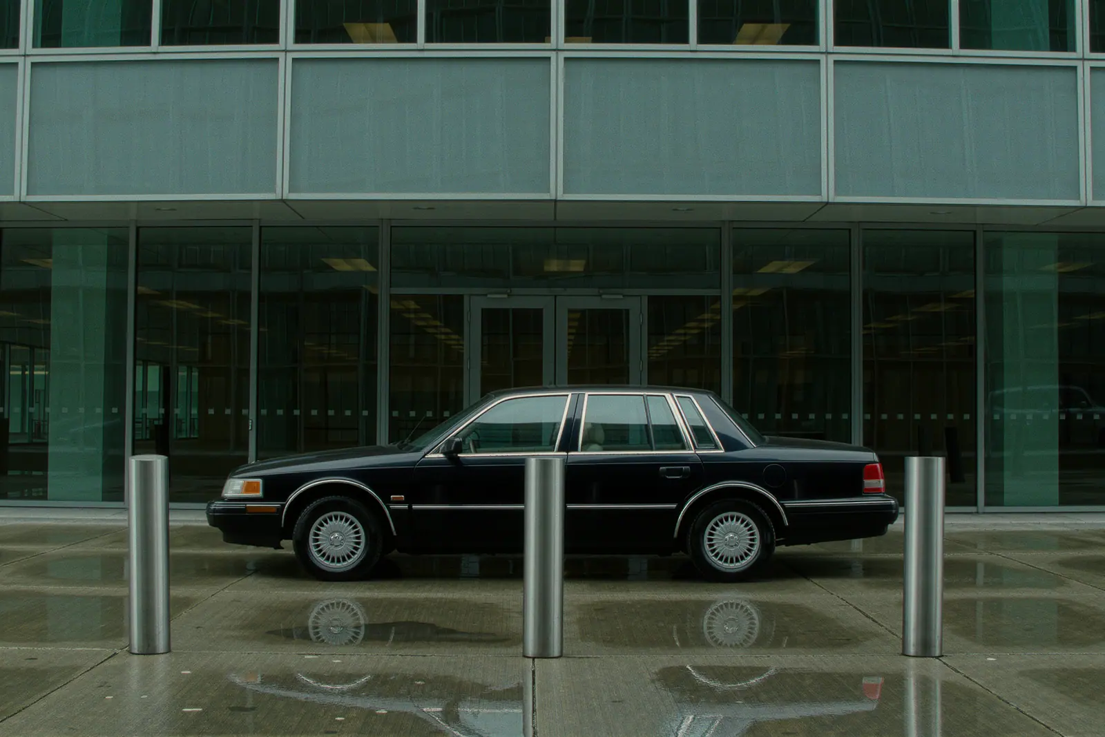 A black unbranded saloon waiting at the entrance of a glass office building with its doors shut and nobody around, a bollard and wet paving slabs in the foreground.