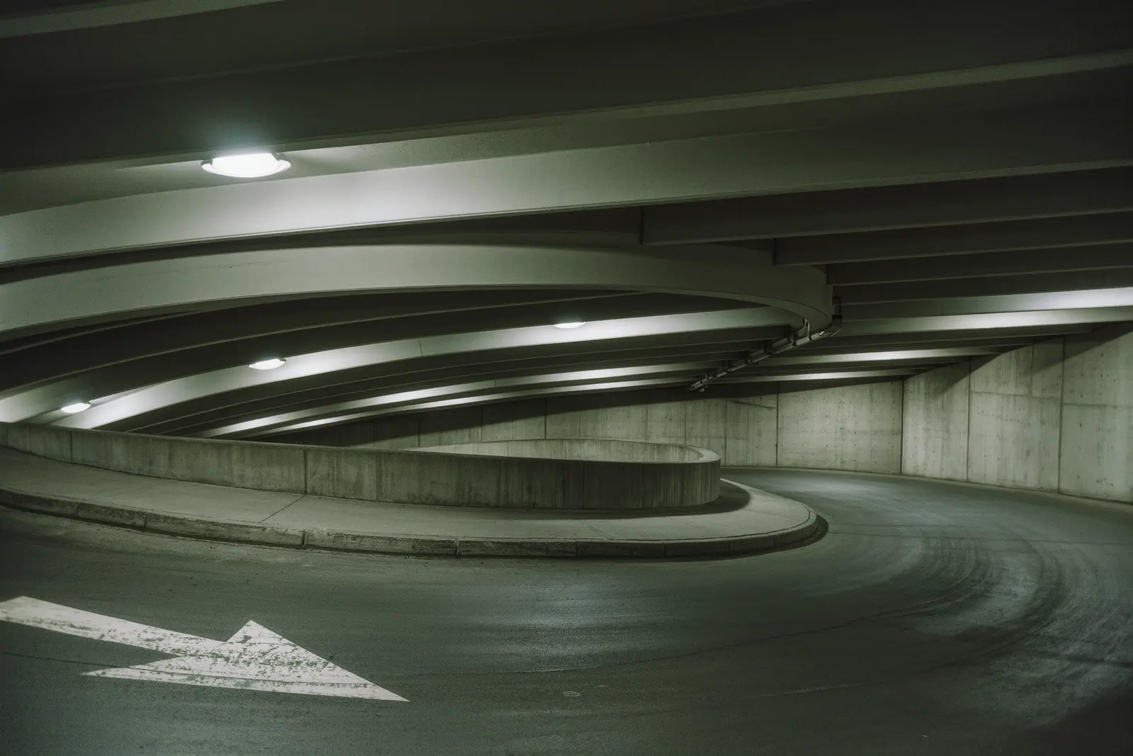 A concrete exit ramp spiralling down out of an airport parking deck, a painted directional arrow and a scuffed kerb in the foreground under low ceiling lighting.