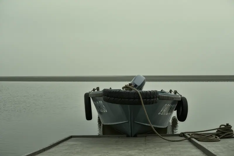 A small open river boat tied alongside a pontoon with fenders hanging over the side, flat grey water and an empty bank behind.