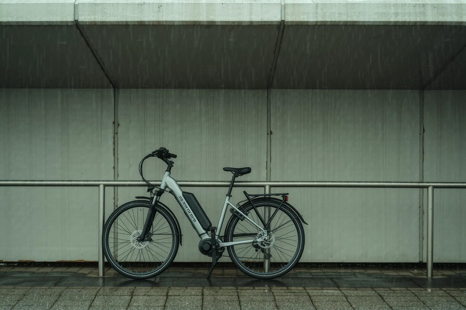 Rental e-bikes locked in a steel rack outside a station entrance, the front wheel and battery of the nearest bike in the foreground on wet paving.